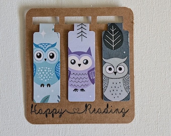 Può includere: Un set di tre segnalibri magnetici rettangolari fissati su un cartoncino marrone con il testo Happy Reading. I segnalibri presentano illustrazioni di gufi in blu, viola e grigio, decorati con motivi di foglie, stelle e alberi.
