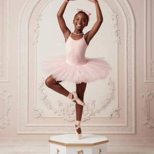 Op de afbeelding: Een jonge balletdanseres in een roze turnpakje en tutu balanceert op spitzen op een witte, goudkleurige muziekdoos. Ze draagt een bijpassende haarband en parelketting, armen sierlijk gebogen. De achtergrond heeft een decoratieve, sierlijke muur.