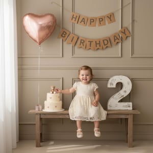 Puede incluir: Una niña con un vestido de encaje blanco está sentada en un banco de madera con una tarta de cumpleaños de dos pisos y un gran número 2. Un globo en forma de corazón flota arriba, y una pancarta dice "Happy Birthday."