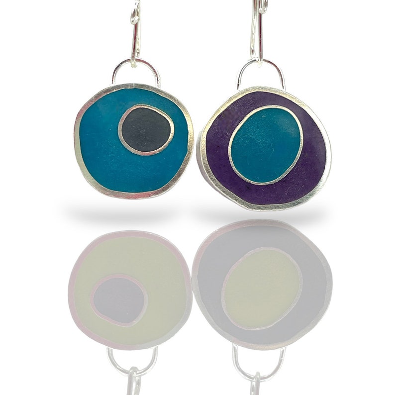 May include: A pair of silver dangle earrings with a teal and purple resin design. The earrings have a round shape with a smaller circle inside. One earring has a black circle inside the teal circle, while the other earring has a teal circle inside the purple circle.