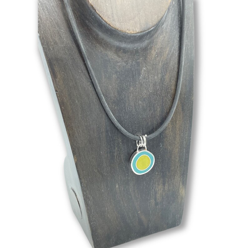 May include: A black cord necklace with a silver pendant featuring a yellow circle surrounded by a turquoise ring.