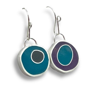 May include: A pair of silver earrings with round resin inlays. One earring has a teal blue inlay with a black circle in the center. The other earring has a purple inlay with a teal blue circle in the center.