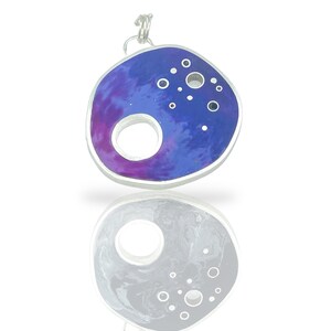 May include: A silver pendant with a purple and blue resin inlay. The pendant has a large circle cut out of the center and is decorated with small circles.