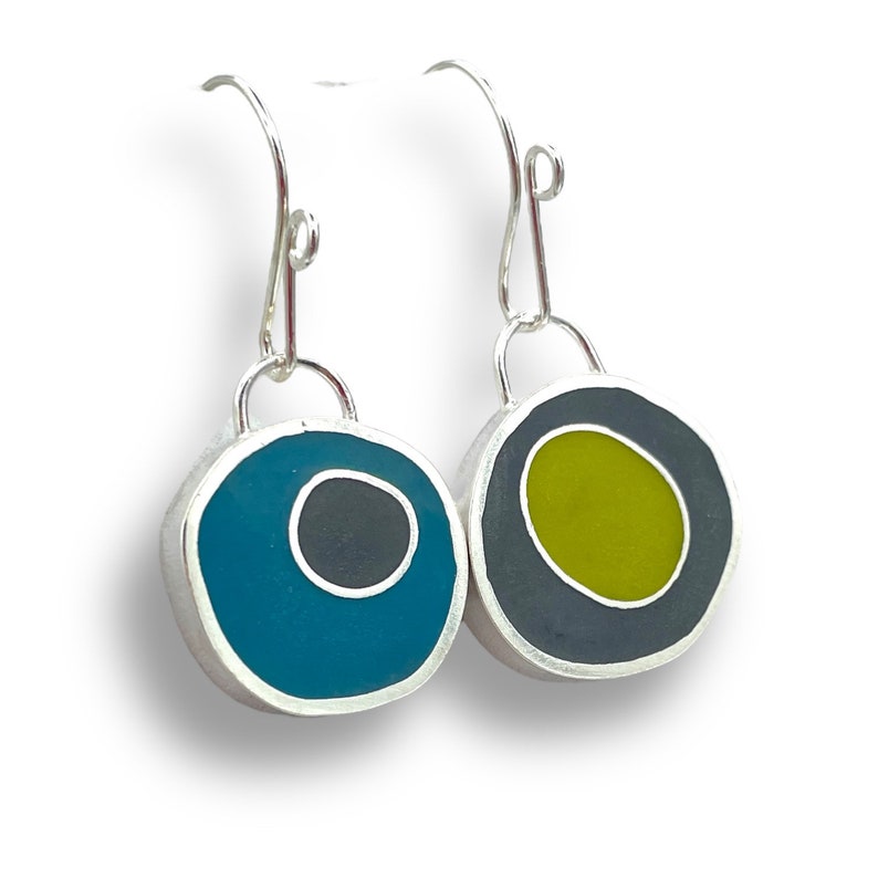 May include: A pair of silver dangle earrings with round resin insets. One earring has a teal blue circle with a black circle inside. The other earring has a gray circle with a yellow circle inside.