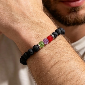 May include: A beaded bracelet featuring black porous lava stone beads, accented by three smooth, translucent spherical beads in green, purple, and red, separated by small silver-toned metal spacers, arranged in a single strand on an elastic cord.