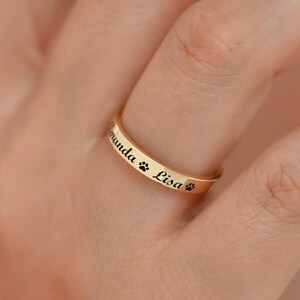 May include: A gold-colored ring with the names "Amanda" and "Lisa" engraved in black, along with paw print symbols. The ring is worn on a finger, showcasing its polished surface and personalized design.