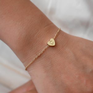 May include: A delicate gold bracelet featuring a small heart charm with the letter "C" engraved on it. The bracelet is a fine chain, and the heart charm is centered. The bracelet is worn on a wrist.