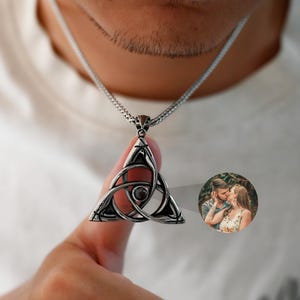 Puede incluir: Collar con colgante plateado con un diseño de nudo celta dentro de un marco triangular. El colgante tiene una pequeña piedra oscura en el centro. También es visible una pequeña imagen circular de una pareja.