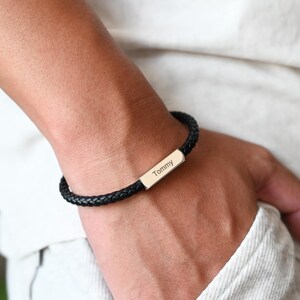 May include: A black braided leather bracelet featuring a rectangular silver plate engraved with the name "Tommy". The bracelet is displayed on a wrist, highlighting the woven texture and personalised detail.
