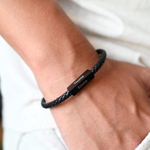 May include: Black braided leather bracelet with a rectangular black metal clasp. The clasp is engraved with the names "Thoma" and "Emma" in white lettering. The bracelet is worn on a wrist.
