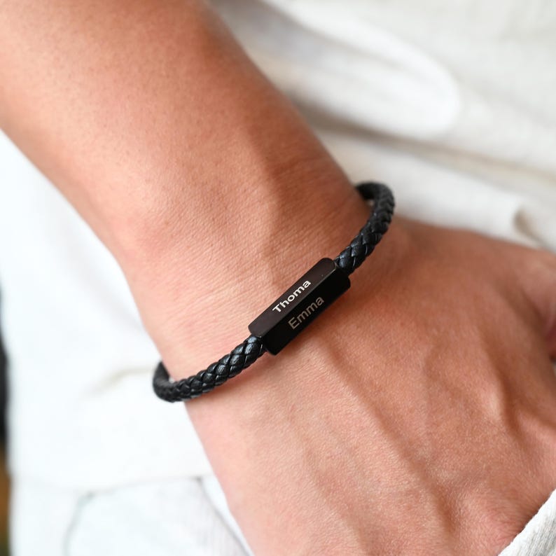 May include: Black braided leather bracelet with a rectangular black metal clasp. The clasp is engraved with the names "Thoma" and "Emma" in white lettering. The bracelet is worn on a wrist.