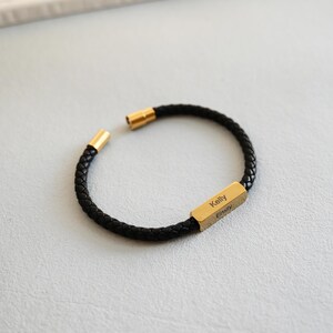 May include: Black braided leather bracelet with gold clasps and a rectangular gold bar engraved with the names "Kelly" and "Emily". The bracelet is displayed on a light grey surface.
