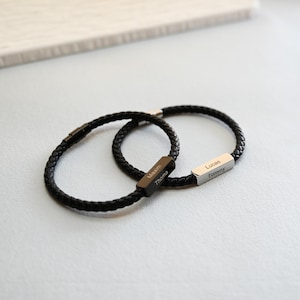May include: Two black braided leather bracelets. One bracelet has a black rectangular clasp engraved with the names "Maxim" and "Thoma". The other bracelet has a silver rectangular clasp engraved with the names "Lucas" and "Tommy".