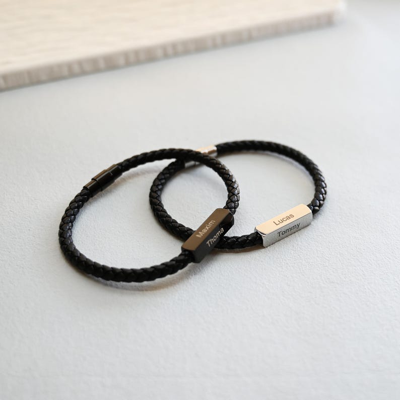 May include: Two black braided leather bracelets. One bracelet has a black rectangular clasp engraved with the names "Maxim" and "Thoma". The other bracelet has a silver rectangular clasp engraved with the names "Lucas" and "Tommy".