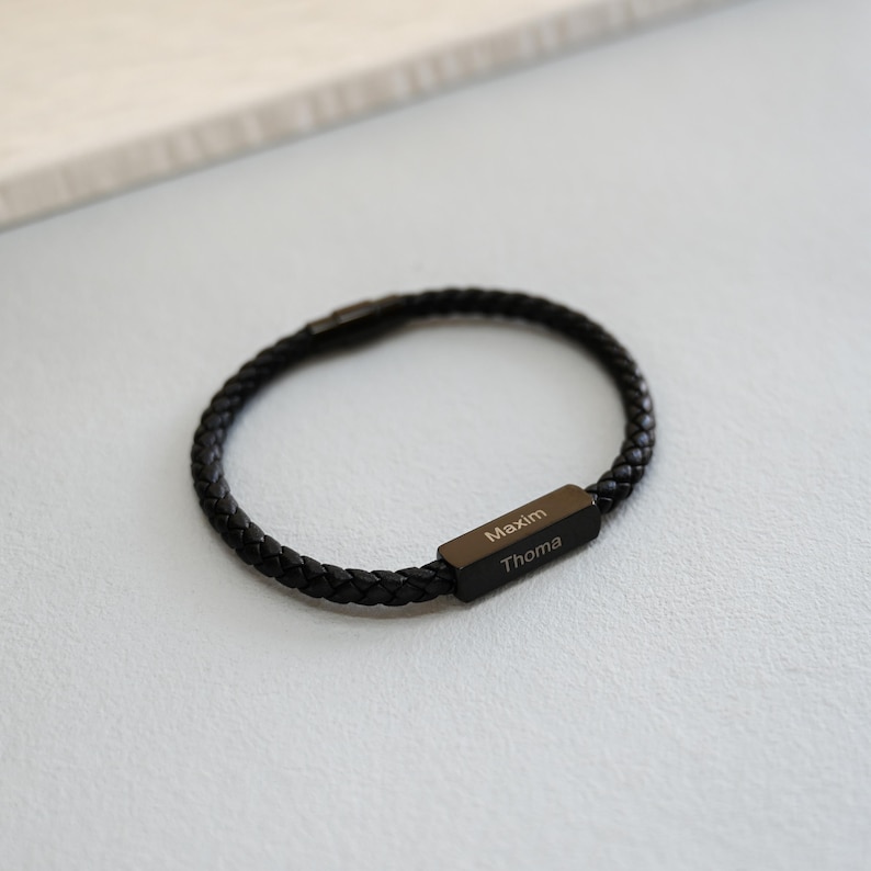 May include: Black braided leather bracelet with a rectangular clasp. The clasp is engraved with the names "Maxim" and "Thoma". The bracelet is a stylish accessory.