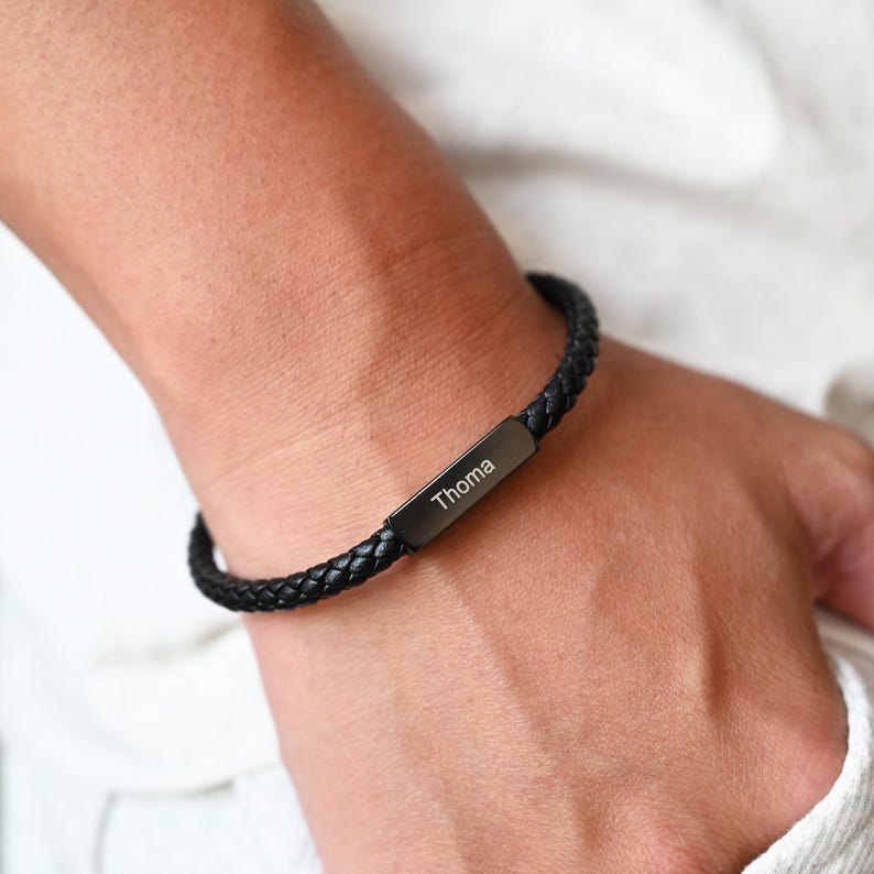 May include: Black braided leather bracelet with a rectangular metal clasp. The clasp is engraved with the name "Thoma" in white. The bracelet is worn on a wrist.