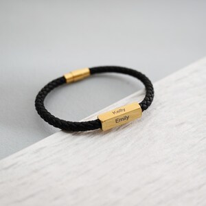 May include: Black braided leather bracelet with gold-coloured rectangular clasp. The clasp is engraved with the names "Kelly" and "Emily". The bracelet is on a light grey and white surface.