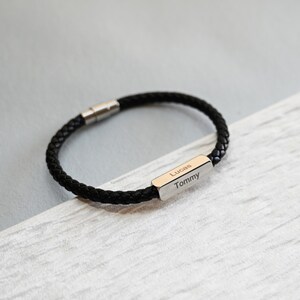May include: Black braided leather bracelet with a silver clasp. The bracelet has a rectangular silver plate engraved with the names "Lucas" and "Tommy". A fashionable accessory.