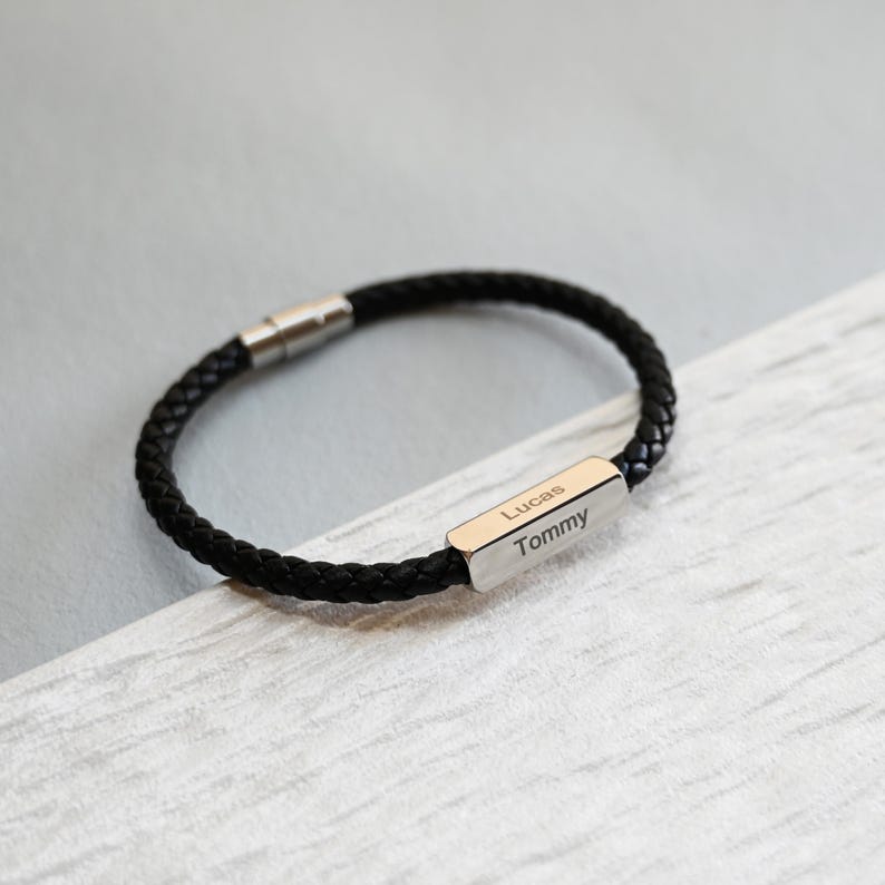 May include: Black braided leather bracelet with a silver clasp. The bracelet has a rectangular silver plate engraved with the names "Lucas" and "Tommy". A fashionable accessory.