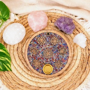 Könnte beinhalten: Ein rundes Holztablett mit einer zentralen Harzeinlage, gefüllt mit bunten Schottersteinen und sieben lasergeschnittenen Chakra-Symbolen aus Holz. Ein kleiner goldfarbener Metallkerzenhalter ist unten eingelassen, umgeben von Kristallen.