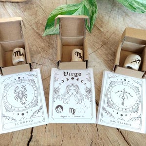 Puede incluir: Tres pequeñas cajas de cartón abiertas contienen cada una un objeto cilíndrico blanquecino con un símbolo del zodiaco. Cada caja se asienta sobre una tarjeta blanca con una ilustración y el nombre del zodiaco: Escorpio, Virgo y Sagitario. El fondo es una superficie de madera.