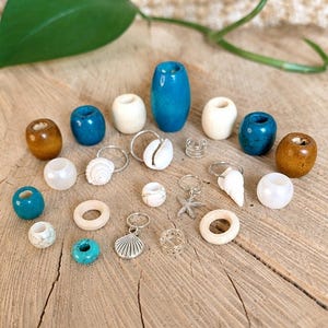 Könnte beinhalten: Eine Sammlung verschiedener Haaraccessoires, darunter blaue, braune und weiße Holzperlen, silberfarbene Metallringe sowie Anhänger in Form von Muscheln und einem Seestern. Das Set bietet verschiedene Texturen und Oberflächen für das Haarstyling.