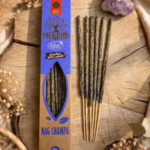 Könnte beinhalten: Eine rechteckige blau-braune Pappschachtel mit Herbio Nag Champa Räucherstäbchen und einer 20g Gewichtsangabe. Die Schachtel zeigt ein Baumlogo, eine Ecocert-Zertifizierung und ein Sichtfenster. Fünf dünne, texturierte, dunkelgrüne Räucherstäbchen.