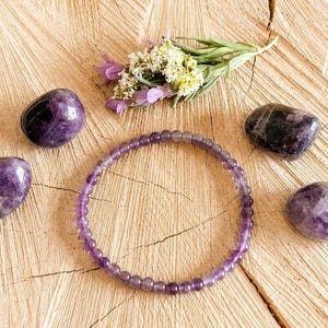 May include: An amethyst beaded bracelet and several polished amethyst stones are arranged on a wooden surface. A small sprig of lavender and white flowers adds a natural accent to the composition. The stones vary in size and shade of purple.