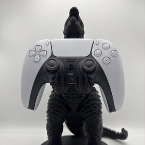 以下が含まれることがあります： 白いPlayStation 5のコントローラーを持った黒いゴジラ像。像の台座は黒で、「GODZILLA」という文字が白で書かれている。
