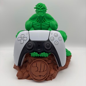 Könnte beinhalten: Eine grüne Hulk-Figur hält einen weißen und schwarzen PlayStation 5-Controller. Der Controller liegt auf einem braunen Sockel mit einem braunen Faustsymbol.