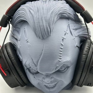 Könnte beinhalten: Ein 3D-gedruckter Kopf einer gruseligen Puppe mit genähten Nähten und einem breiten, zahnigen Grinsen, der einen schwarzen Over-Ear-Kopfhörer mit roten Akzenten trägt.