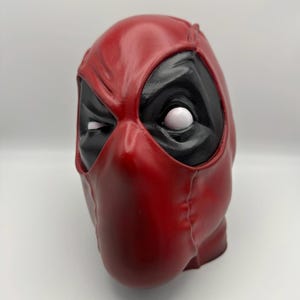 May include: A red and black Deadpool mask with a white eye. The mask is made of a textured material and has a stitched seam down the centre.