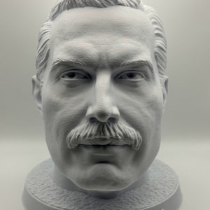 Può includere: Un busto bianco di Freddie Mercury, il cantante principale dei Queen, stampato in 3D. Il busto è su una base rotonda con il nome "Freddie Mercury" inciso su di essa.