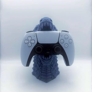 Könnte beinhalten: Ein hellblauer PlayStation 5-Controller, der auf einem blauen Monster-Ständer ruht. Der Controller hat schwarze Akzente, und der Ständer zeigt ein detailliertes Monsterkopf-Design mit scharfen Zähnen. Der Hintergrund ist ein weiches, weißes Farbverlauf.