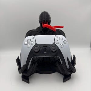 Könnte beinhalten: Eine schwarz-rote 3D-gedruckte Venom-Figur hält einen weißen PlayStation 5-Controller. Der Controller liegt in den Händen der Figur.