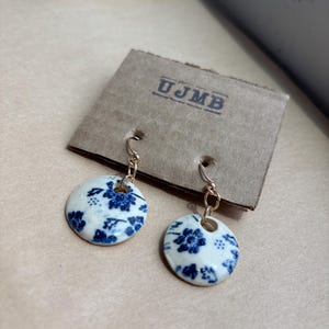 Puede incluir: Pendientes redondos con un diseño floral azul y blanco, suspendidos de ganchos dorados. Los pendientes se exhiben en un respaldo de cartón con las letras "UJMB". El fondo es una superficie marrón claro.