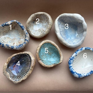 以下が含まれることがあります： 手作りの貝殻型セラミックボウル6個。各ボウルは、青、ターコイズ、ニュートラルな色合いなど、ユニークなデザインとカラーパレットを持っています。ボウルはニュートラルな背景に配置され、有機的な形と質感を際立たせています。