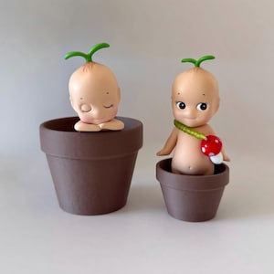 May include: Two small, brown terracotta pots each hold a small, nude baby-like figure with a green sprout on its head. One figure has closed eyes, the other has open eyes and a mushroom accessory. The figures are whimsical and charming.