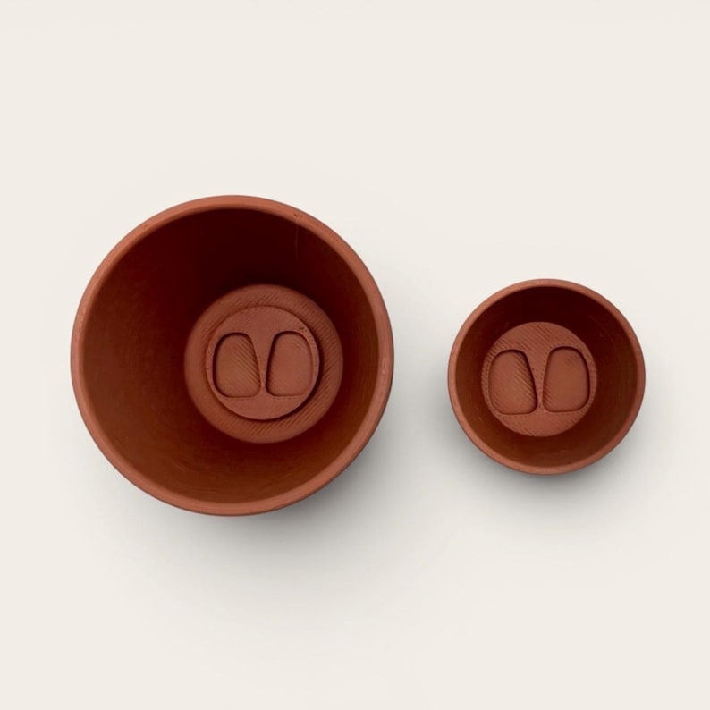 May include: Two terracotta-colored ceramic bowls. The larger bowl is on the left, and the smaller bowl is on the right. Both bowls have a circular base with a raised, oval-shaped design in the center. The bowls are set against a white background.