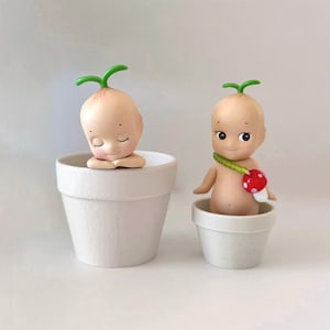 May include: Two small, light-skinned baby-like figurines with green sprout-like hair, sitting in white pots. One figurine has closed eyes and clasped hands. The other has open eyes and a mushroom necklace. The figurines are a unique decorative item.