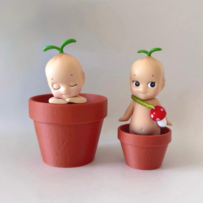 May include: Two small, terracotta-colored flower pots each hold a unique doll. One doll has closed eyes and a green sprout on its head. The other doll has open eyes, a mushroom accessory, and a green sprout.