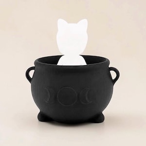 May include: A matte black cauldron with two side handles and three small feet. The front features an embossed triple moon symbol. A white, flat, cat-shaped silhouette is positioned inside the cauldron, extending upward from the opening.