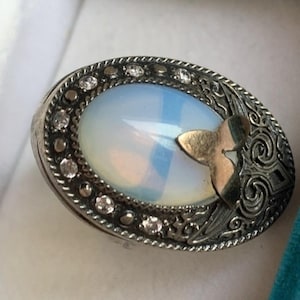 May include: Oval-shaped silver ring with a large, opalescent stone. The ring features a gold-colored accent and is embellished with small, clear gemstones around the edge. The design includes intricate metalwork and a decorative pattern.