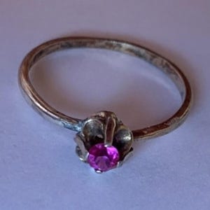 May include: A silver-toned metal ring featuring a thin, smooth band and a prong setting that holds a single, round, vibrant pink gemstone. The metal surface shows a slightly weathered, matte finish throughout the band and the setting.