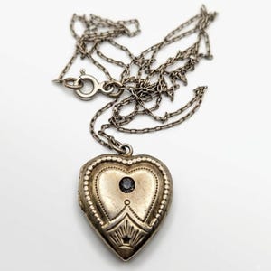 May include: A silver-toned heart-shaped locket pendant suspended from a matching delicate link chain. The locket features an embossed decorative border, a sunburst pattern at the base, and a single small dark circular stone set in the center.