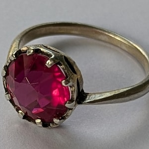 May include: A silver-toned ring featuring a round, faceted, deep pink gemstone set in a prong setting. The metal band is smooth and tapers slightly toward the back, while the stone is held securely by multiple small metal prongs around the edge.