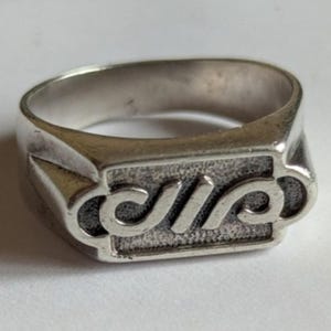 May include: A silver-toned metal ring featuring a rectangular face with a textured, recessed background. The center of the face displays a raised, stylized, continuous looping design that resembles a cursive letter or an abstract symbol.