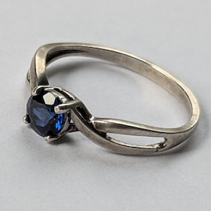 May include: A silver-toned ring featuring a single round-cut dark blue gemstone held in a four-prong setting. The band has a split, crossover design that tapers toward the back, creating a smooth, polished metallic appearance throughout.