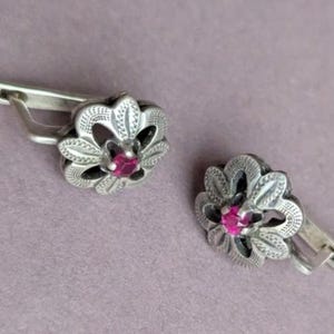 May include: A pair of silver-toned earrings featuring a flower-shaped design with intricate etched detailing on the petals and a small, round, vibrant pink gemstone set securely in the centre of each piece, complete with a hinged lever-back closure.