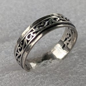 May include: A silver ring with an intricate openwork design. The ring features a repeating pattern of swirling shapes and lines. The metal has a polished finish, and the ring is set against a textured, white background.